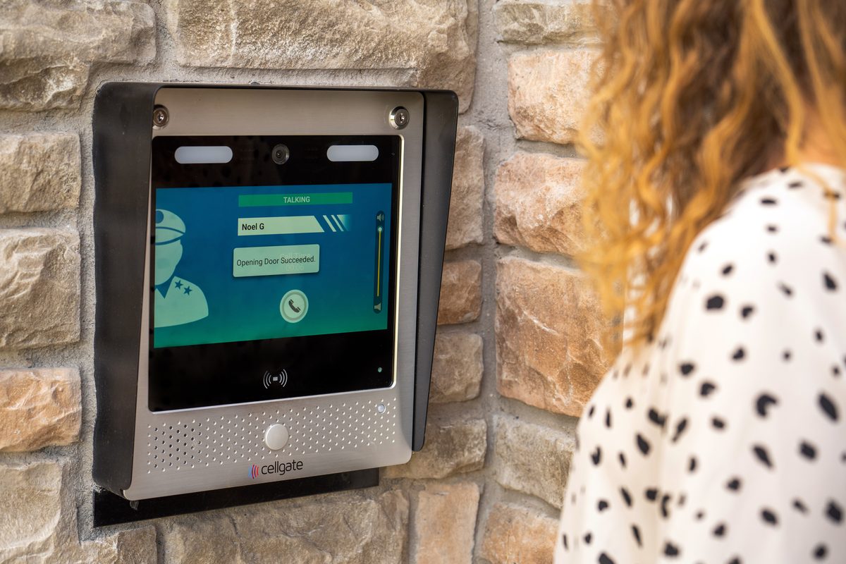 Woman placing a video call from a CellGate Watchman WXL touchscreen mounted in a stone wall at a community entrance